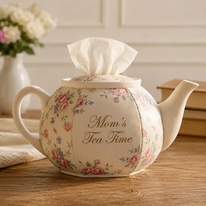 Puede incluir: Una caja para pañuelos de cerámica con forma de tetera, con base color crema y un estampado floral de rosas rosas y hojas azules. El texto "Mom's Tea Time" está impreso en letra cursiva marrón en el frente. Un pañuelo blanco sobresale por arriba.