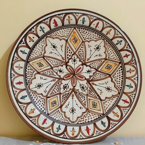 Puede incluir: Un plato redondo de cerámica con un patrón geométrico y floral pintado a mano en tonos marrón, amarillo, rojo y verde sobre un fondo crema. El diseño incluye un motivo de estrella central, líneas intrincadas y un borde festoneado.