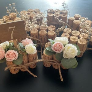 Può includere: Disposizione di tappi di sughero, alcuni legati con spago, e sormontati da fiori artificiali bianchi e rosa. Segnaposto in legno con numeri sono posizionati tra i tappi. Il tema è rustico e decorativo.