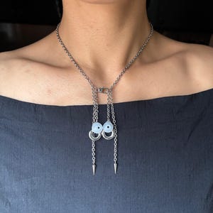 Op de afbeelding: Zilverkleurige ketting met een ketting en een hanger met twee lichtblauwe kralen en zilveren ringen. De hanger hangt aan een ketting met twee zilveren kegelvormige bedels. De ketting wordt door een persoon gedragen.