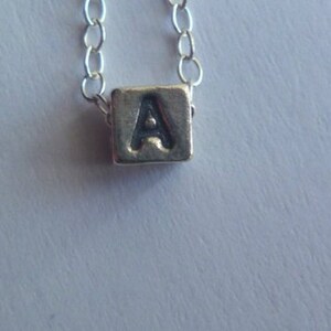 May include: Silver necklace featuring a small, square pendant with the letter "A" engraved on it. The pendant is suspended from a delicate chain with small, oval links. The background is a light, neutral color.