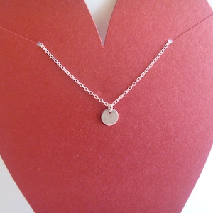 May include: A silver chain necklace with a small, round silver pendant. The necklace is displayed on a red heart-shaped background.
