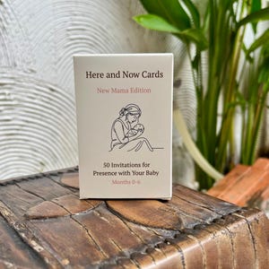 May include: A rectangular white card deck box titled Here and Now Cards New Mama Edition. The front features a black line drawing of a person holding a baby, with the text 50 Invitations for Presence with Your Baby Months 0-6 printed below.