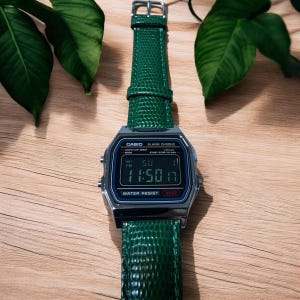 May include: A digital wristwatch with a silver-tone rectangular case and a textured dark green leather strap. The watch face features a black digital display showing the time, date, and day, with the text CASIO, ALARM CHRONO, and WATER RESIST.