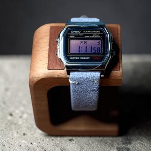 May include: A Casio digital watch with a silver-tone rectangular case and a light blue suede strap. The watch face displays the text CASIO, ALARM CHRONO, LITHIUM, WATER RESIST, and WR. The digital screen shows the time 1:11 and the day FR 3.