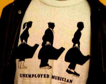 Op de afbeelding: Wit t-shirt met een zwarte afbeelding van drie figuren die instrumentenkoffers dragen. De tekst "UNEMPLOYED MUSICIAN" staat eronder. Een zwarte leren jas is over het shirt gedrapeerd.