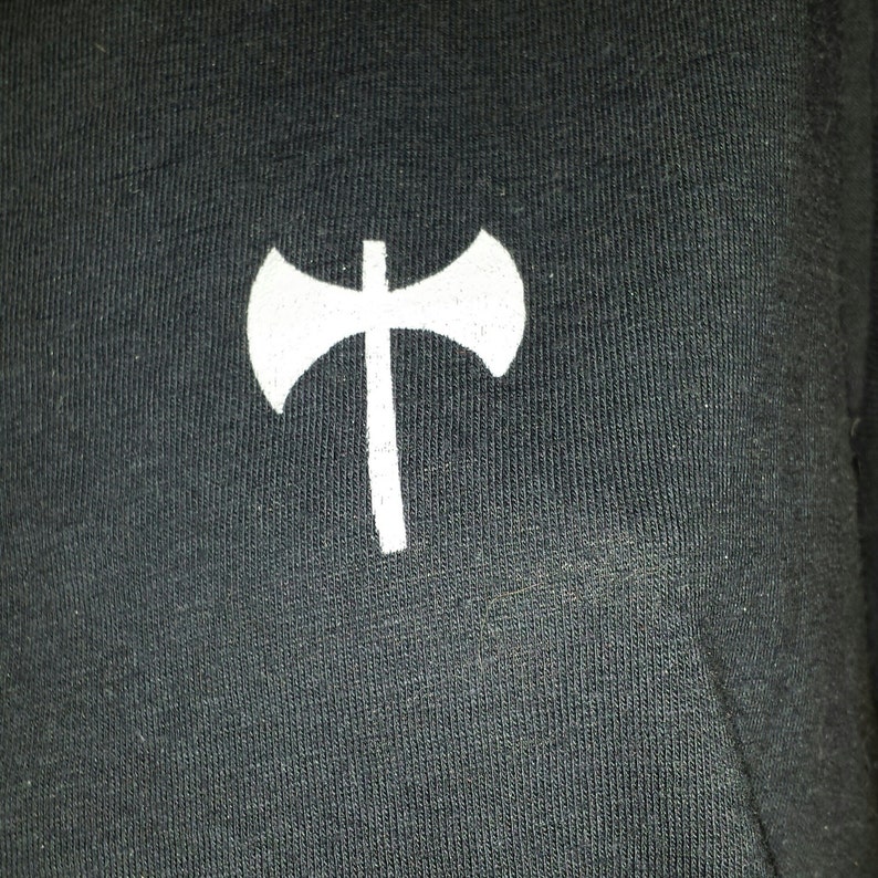 K&ouml;nnte beinhalten: Ein wei&szlig;es Axt-Symbol auf einem schwarzen Stoffhintergrund.