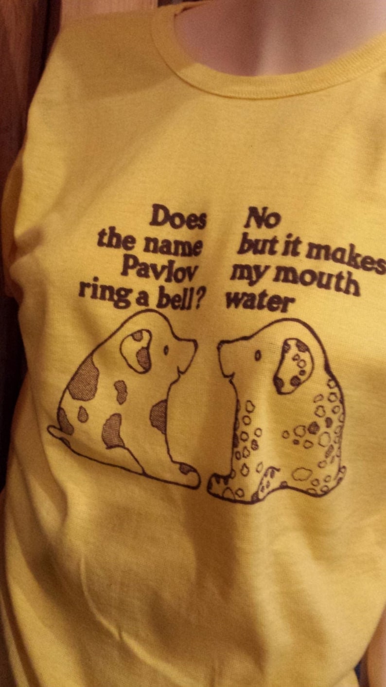 May include: A yellow t-shirt with brown text that reads 'Does the name Pavlov ring a bell? No but it makes my mouth water.' The text is accompanied by two cartoon dogs with brown spots.