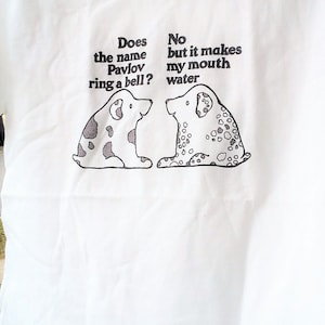 May include: White t-shirt with a black and white illustration of two dogs. The text on the shirt reads "Does the name Pavlov ring a bell?" and "No but it makes my mouth water."