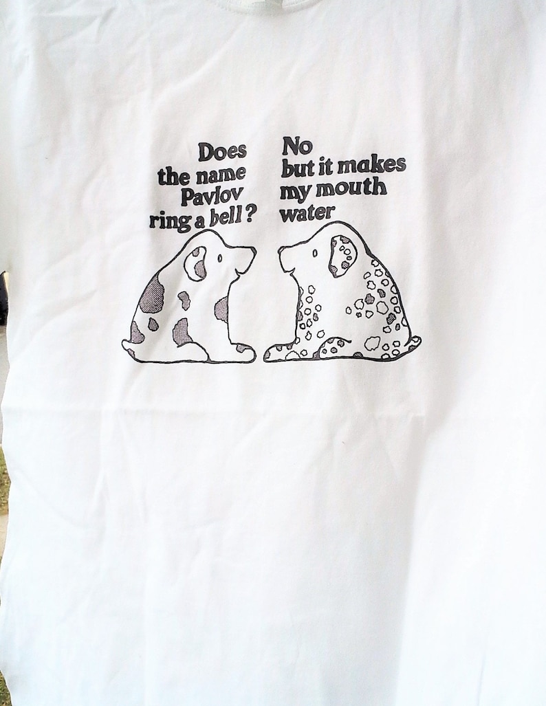 May include: White t-shirt with a black and white illustration of two dogs. The text on the shirt reads "Does the name Pavlov ring a bell?" and "No but it makes my mouth water."