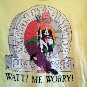 Peut inclure: T-shirt jaune avec une illustration de dessin animé d'un homme en robe de juge chevauchant un bison. L'illustration est entourée d'un cercle avec le texte "Department of Interior 1849-1981". Le texte "Watt? Me Worry!" est imprimé sous l'illustration.