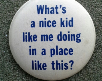 Puede incluir: Una chapa redonda de color blanco con el texto en azul What's a nice kid like me doing in a place like this? El accesorio circular tiene una superficie lisa y letras claras y en negrita centradas sobre el fondo blanco.