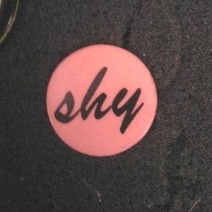 Op de afbeelding: Een ronde, lichtroze knop met het woord "shy" in zwart cursief schrift. De knop staat tegen een donkere, getextureerde achtergrond. Een eenvoudig, maar opvallend ontwerp.