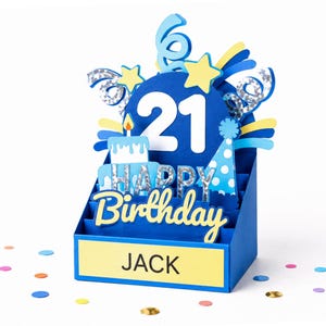 May include: A blue 3D pop-up birthday card featuring the number 21 in white, a cake, a party hat, and stars. The card displays the text HAPPY BIRTHDAY and the name JACK on a yellow rectangular base, accented with silver holographic details.