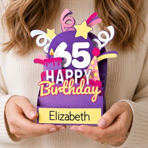 May include: A purple 3D pop-up birthday card featuring the number 65, a lit candle, a party hat, and colorful streamers. The text reads Happy Birthday and Elizabeth on a yellow rectangular base. The design includes yellow stars and pink accents.