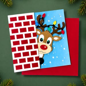 May include: A greeting card featuring a brown reindeer with a red nose peeking from behind a white and red brick wall pattern. The reindeer has brown antlers wrapped in a string of colourful Christmas lights against a light blue snowy background.