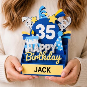 May include: A three-dimensional blue birthday card featuring the number 35, a cake, and a party hat. The card displays the text HAPPY BIRTHDAY in silver glitter and the name JACK in black letters on a yellow rectangular base with star accents.
