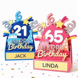 Two 3D layered paper birthday cards. The blue card features the number 21, a cake, and the name JACK. The pink card features the number 65, a cake, and the name LINDA. Both cards display the text HAPPY BIRTHDAY with silver accents.