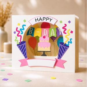 May include: A white 3D pop-up greeting card featuring a circular cutout with a layered paper design. Inside, a two-tier pink cake sits on a stand beneath a colourful "B-DAY" banner and balloons. Two purple party poppers with streamers flank the centre.