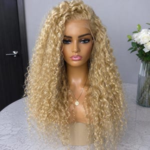 Puede incluir: Una peluca larga y rizada de color rubio sobre una cabeza de maniquí. La peluca presenta una construcción frontal de encaje con una línea de cabello natural, rizos en espiral apretados y una paleta de colores rubios con reflejos claros.
