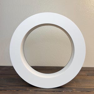 May include: A circular white frame with a thick border and a hollow center. The object has a smooth, matte finish and a flat front surface, creating a simple geometric ring shape that stands upright on its own edge.