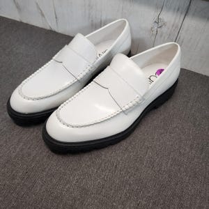 May include: A pair of white loafers featuring a smooth, glossy finish, a classic penny slot strap across the vamp, and a thick, black lugged rubber sole. The interior of the shoe shows a partial brand logo and a small circular purple sticker.