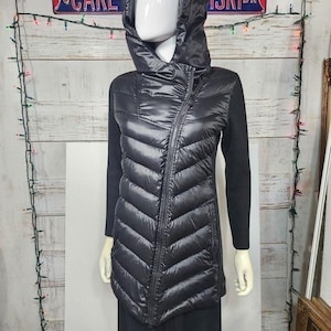 Könnte beinhalten: Eine schwarze Langarmjacke mit gestepptem, glänzendem Vorderteil im Daunenjacken-Stil, asymmetrischem Reißverschluss, Kapuze und Ärmeln aus schwarzem Strickstoff. Das Kleidungsstück hat eine taillierte Passform und einen hohen Stehkragen.