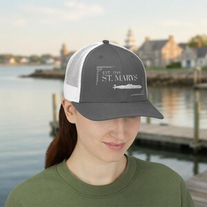 May include: A charcoal grey trucker hat with a white mesh back. The front panel features a white graphic print with the text "EST. 1792 ST. MARYS" above a line drawing of a submarine, framed by thin white geometric corner accents.