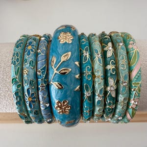 May include: A set of nine bangle bracelets in varying shades of blue, teal, and green. The bracelets feature gold-tone floral and leaf patterns, some with inlaid clear crystals. The central bangle is wider than the others and has a marbled finish.