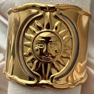 Pode incluir: Uma pulseira larga de tom dourado polido com um motivo central de sol em relevo apresentando um rosto humano. O sol é rodeado por raios e emoldurado por uma borda circular, sobre uma banda de metal lisa e curva com bordas onduladas.