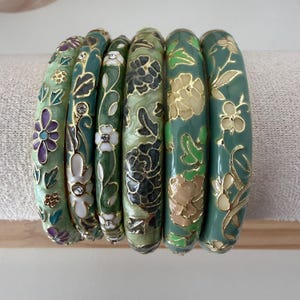 May include: A set of six green bangle bracelets featuring various floral patterns in shades of purple, white, and gold. Each bracelet is decorated with enamel detailing and some include small embedded clear crystals along the floral designs.