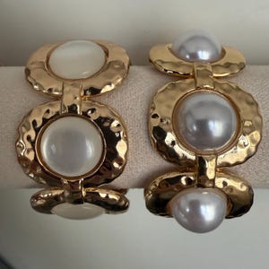 May include: Two link bracelets featuring a hammered gold-tone metal finish. The left bracelet displays smooth, opaque white circular cabochons, while the right bracelet features lustrous, iridescent silver-toned spherical faux pearls set within each circular link.