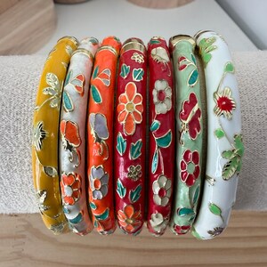 May include: A set of seven cylindrical bangles featuring colorful enamel finishes in yellow, white, orange, red, and light green. Each bangle is decorated with gold-toned metal floral patterns and leaves that wrap around the entire surface.