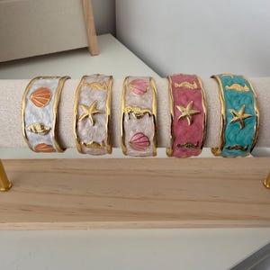 Puede incluir: Cinco brazaletes de metal en tono dorado con borde ondulado y acabado de esmalte marmoleado en blanco, rosa claro y turquesa. Cada brazalete está decorado con dijes dorados en relieve con motivos marinos, incluyendo estrellas de mar y conchas.