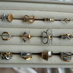 Pode incluir: Uma coleção de doze anéis dispostos em três filas, apresentando vários designs, incluindo formas de estrela-do-mar, concha, coração, flor e estrela. Os anéis são feitos em tons mistos de ouro e prata, com bandas metálicas lisas, onduladas e texturizadas.