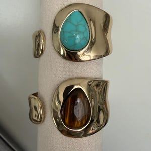 May include: Two gold-toned cuff bracelets with irregular, organic shapes. The top bracelet features a teardrop-shaped turquoise stone with dark veining. The bottom bracelet features a teardrop-shaped brown tiger's eye stone with a chatoyant finish.