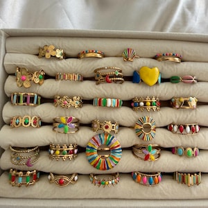 May include: A collection of gold-tone metal rings arranged in rows, featuring various designs including colorful enamel flowers, hearts, moons, stripes, and geometric patterns. Each ring displays vibrant multi-colored accents in a variety of shapes.