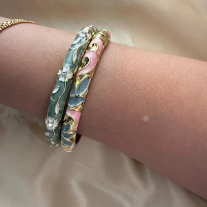 May include: Two gold bangles with floral designs. One bangle is green with white flowers, the other is pink with blue and gold accents. The bracelets are worn on a person's arm.