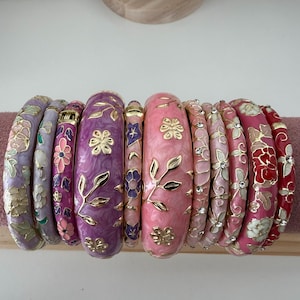May include: A collection of eleven hinged bangle bracelets in varying shades of pink, purple, and red. Each bracelet features gold-tone metal floral and vine motifs, with some designs accented by small clear crystals set into the enamel surfaces.