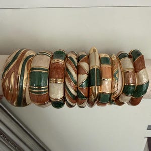 May include: A collection of ten gold-tone metal bangles featuring enamel inlays in shades of cream, forest green, and terracotta. The bracelets display various patterns including stripes, geometric triangles, and solid color-blocked segments.