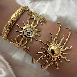 Peut inclure: Trois bracelets dorés. L'un est une manchette large avec des gravures d'étoiles et de lunes et de petites pierres incrustées. Le deuxième est une manchette avec plusieurs motifs de soleil à centre émaillé blanc. Le troisième est un jonc texturé.