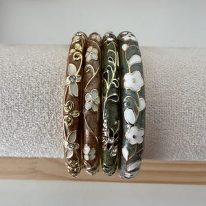 May include: A set of four bangle bracelets featuring floral patterns in shades of brown, green, and white. Each bracelet is accented with gold-tone metal outlines and small clear crystals embedded within the flower designs on the surface.