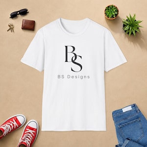 May include: A white short-sleeved t-shirt featuring a black logo with the letters BS and the text BS Designs printed in the center. The shirt is displayed alongside black sunglasses, a brown wallet, keys, two small potted plants, and blue jeans.
