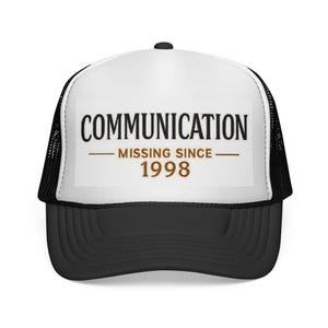 May include: A black and white trucker hat featuring a white front panel with the black text COMMUNICATION and the brown text MISSING SINCE 1998 printed in the center. The cap has a black mesh back, a black brim, and a black button on the top.