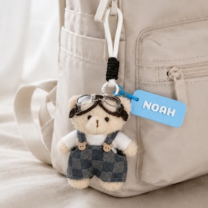 May include: A plush teddy bear keychain wearing aviator goggles and denim overalls. The bear has a white shirt underneath and a blue tag that reads "NOAH". The keychain is attached to a beige backpack.