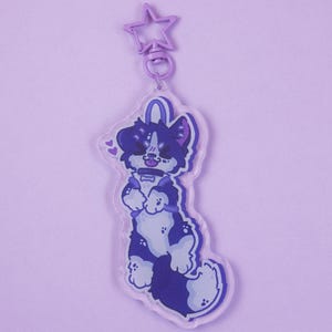 Peut inclure: Un porte-clés en acrylique transparent représentant un chien de dessin animé avec une fourrure violet foncé et lavande clair. Le chien porte un collier avec une médaille en forme d'os et deux petits cœurs près de la tête.