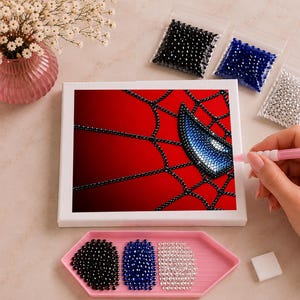 May include: A diamond painting kit featuring a red background with a black web pattern and a blue eye shape. The set includes a white canvas, a pink applicator tool, a pink tray with black, blue, and silver beads, and three small bags of beads.