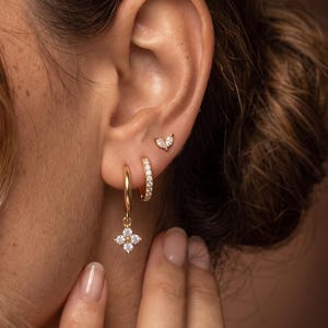 Può includere: Un set di orecchini color oro composto da un piccolo cerchio con un ciondolo a forma di fiore a quattro petali con pietre trasparenti, un cerchio dorato semplice e un minuscolo orecchino a lobo a forma di cuore con una pietra trasparente.