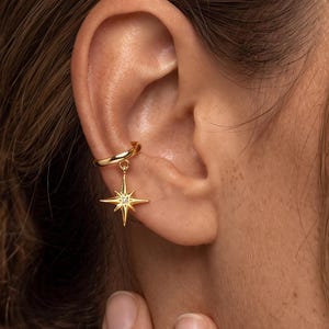 May include: A gold-toned ear cuff featuring a dangling North Star charm. The charm is shaped like a four-pointed star with elongated rays and a small, clear, round stone set in the center. The ear cuff is a simple, smooth, circular band.