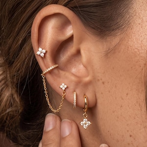 Könnte beinhalten: Ein Set aus goldfarbenen Ohrringen und einer Ohrmanschette mit klaren Steinen. Die Ohrmanschette ist über eine zarte Goldkette mit einem Ohrstecker verbunden. Ein kleiner Creolen-Ohrring mit einem vierblättrigen Blumenanhänger und ein dünner Creolen-Ohrring.